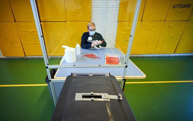 een stembureualid reinigt potloden in een stemlokaal in Oisterwijk. Hij zit achter een kuchscherm, draagt een mondkapje en handschoenen. Op de voorgrond staat een stembus